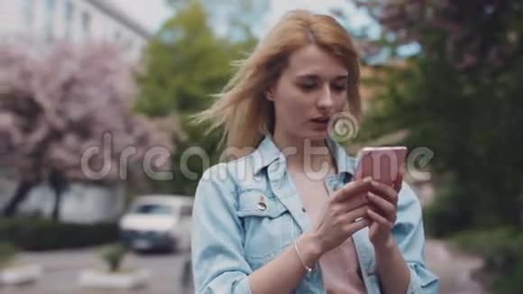 穿着牛仔裤夹克的年轻女士正在城里用她的手机城市建筑背景上的汽车刮风的天气视频的预览图
