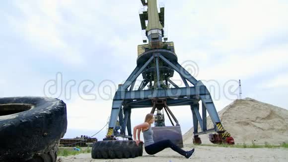 穿着灰色上衣和黑色紧身裤的运动妇女使用大型拖拉机轮进行力量练习做俯卧撑视频的预览图