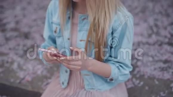 穿着牛仔裤夹克和粉红色连衣裙的漂亮金发女孩在公园里留言秋天的樱花视频的预览图