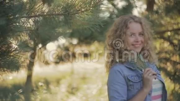 镜头前一个年轻的金发女孩害羞的微笑穿牛仔裤背景上绿色针叶树外面视频的预览图