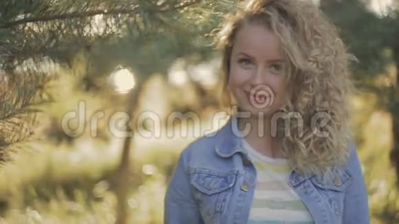 弯弯的金发女孩站在树下阳光明媚背景上的树花园女性肖像视频的预览图
