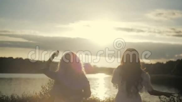 女孩们在湖边的日落前平静下来年轻的雌性跳跃着在灿烂的阳光下挥舞着头发视频的预览图
