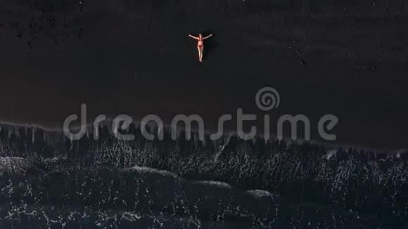 一个穿红色泳衣的女人躺在冲浪线上的黑色海滩上的俯视图加那利特内利岛海岸视频的预览图