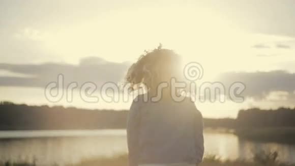 穿着牛仔裤夹克的可爱卷曲的年轻女人沿着湖边跑到灿烂的日落然后转向相机微笑视频的预览图