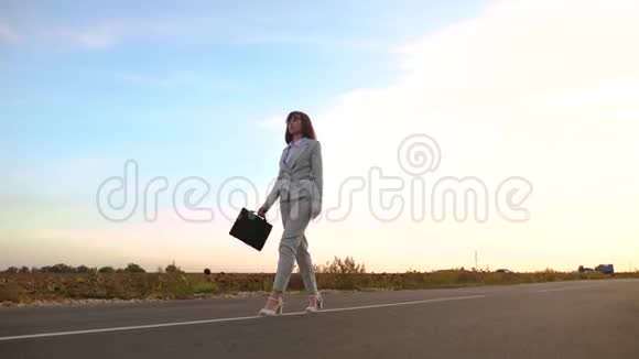 女商人带着一个黑色公文包穿着浅色西装穿着白色高跟鞋沿着沥青路走上面有标记视频的预览图