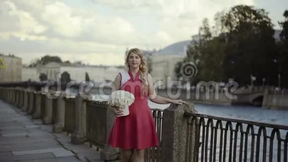 可爱的金发女郎穿着红色连衣裙带着鲜花走在街上视频的预览图