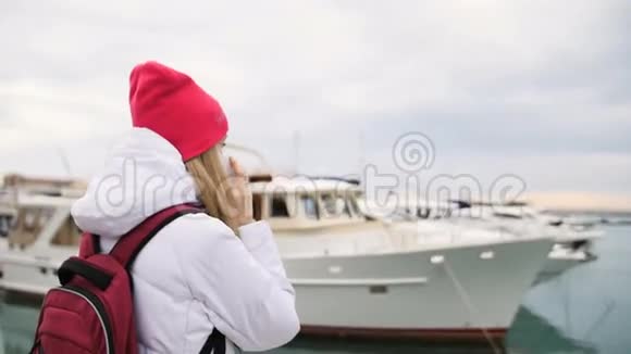 年轻美丽的白种人女孩站在码头上拿着船和手机交谈视频的预览图