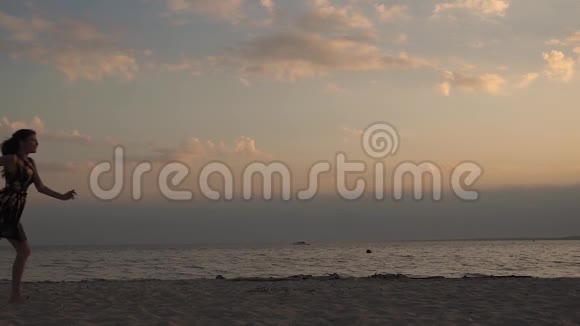 美丽快乐的年轻女孩沿着海滩奔跑快乐地挥动着她的手臂海滩日落时分的年轻女子视频的预览图