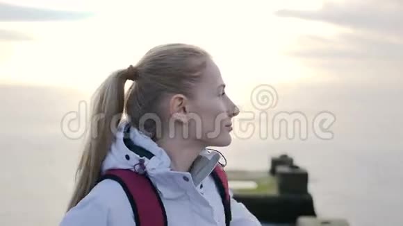 美丽的年轻微笑的女人穿着夹克背着背包在码头上背景是海视频的预览图