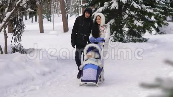 年轻漂亮的穿着衣服的父母走在白雪覆盖的公园里你的孩子坐在婴儿马车上滑雪视频的预览图