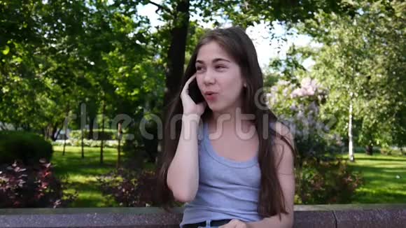 年轻迷人的女孩在长凳上和电话交谈夏日公园微笑高清视频镜头拍摄静态摄像机视频的预览图
