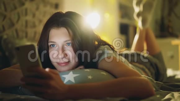 迷人的微笑女性使用智能手机分享社交媒体晚上躺在家里的床上视频的预览图