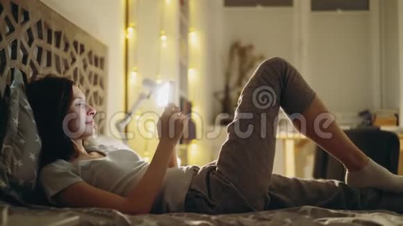 一位年轻的微笑女性她用智能手机分享社交媒体晚上躺在家里的床上视频的预览图