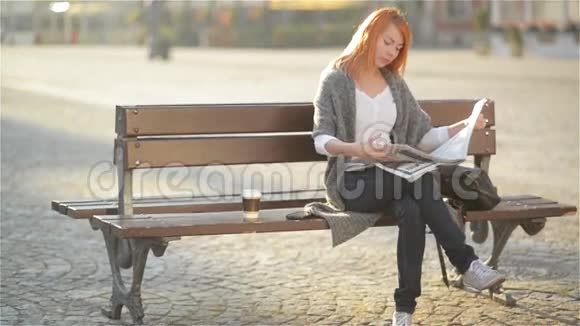 微笑的女人红头发坐在长凳上喝咖啡看报纸阳光明媚的一天视频的预览图