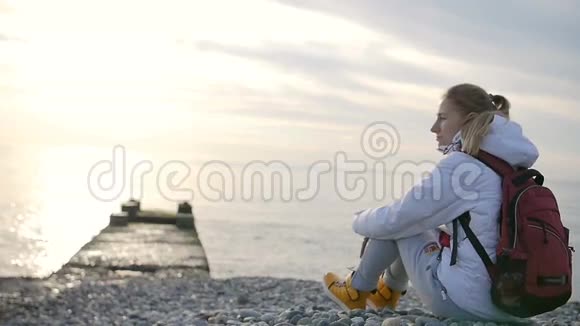 年轻漂亮的女人穿着夹克背着背包日落时坐在海边视频的预览图