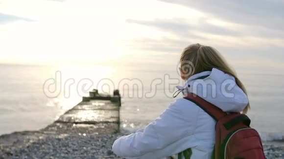 年轻漂亮的女人穿着夹克背着背包日落时坐在海边视频的预览图