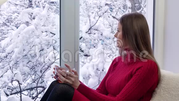 漂亮的红裙子女孩坐在窗台上喝着茶看着窗户冬天在外面视频的预览图