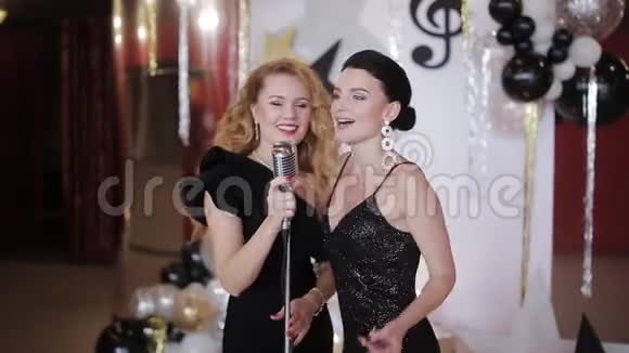 两个身穿晚礼服的迷人女孩对着老式麦克风唱歌在聚会上玩得很开心视频的预览图