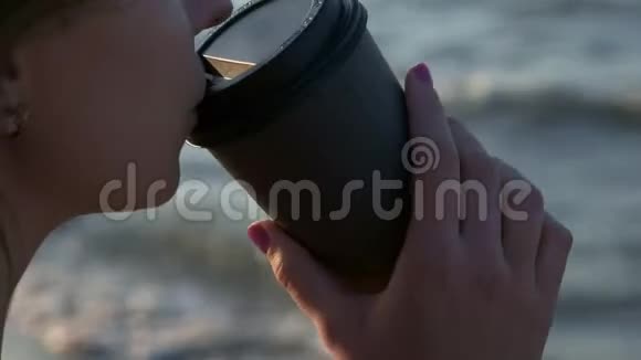 穿着黑色泳衣的女孩在海滩上从纸板玻璃上喝咖啡日落时分在海边或视频的预览图