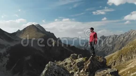 一个留着胡子戴着帽子戴着墨镜的年轻嬉皮士正站在高高的山上的岩石山脊上视频的预览图