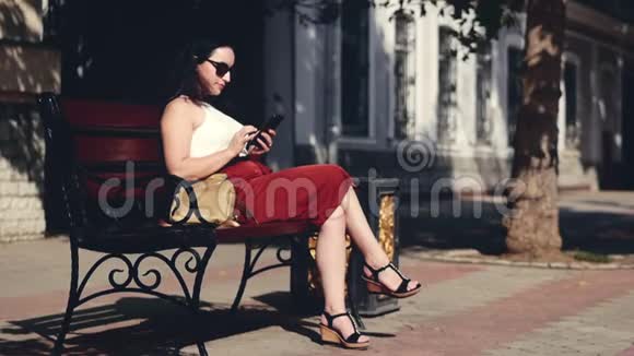 一个漂亮的商界女性一个高加索人的外表使用智能手机技术应用程序坐在长凳上的城市街道上视频的预览图