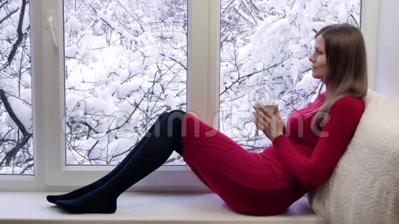 漂亮的红裙子女孩坐在窗台上喝着茶看着窗户冬天在外面视频的预览图