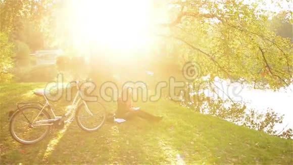 美丽的女孩在秋天公园看书放松坐在湖边的草地上骑着自行车晴天视频的预览图