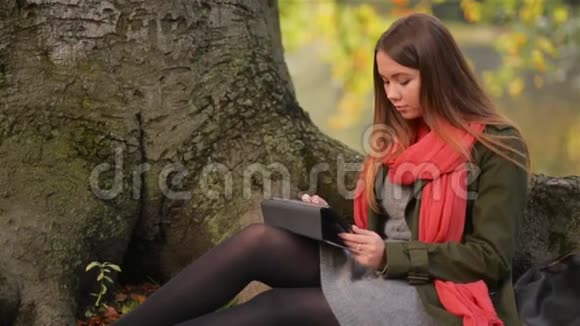 美丽的学生女孩使用平板电脑户外坐在湖边的树上视频的预览图