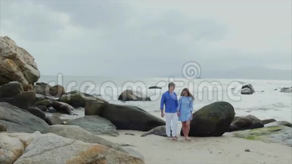 一对年轻幸福的恋人穿着蓝色衣服牵着手微笑着走在海边视频的预览图