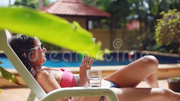 年轻漂亮的女人戴着太阳镜和比基尼用平板电脑躺在游泳池的日光浴床上1920x1080视频的预览图