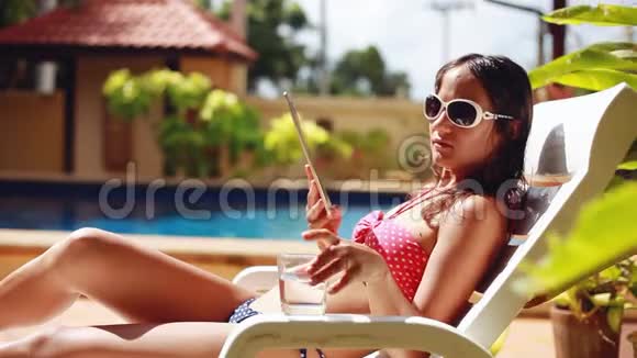 年轻的黑发女人戴着太阳镜和比基尼用平板电脑躺在游泳池旁的日光浴床上喝水1920x1080视频的预览图