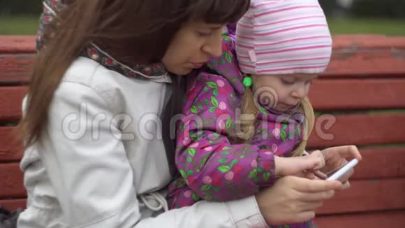 母女俩坐在户外的长凳上玩智能手机视频的预览图
