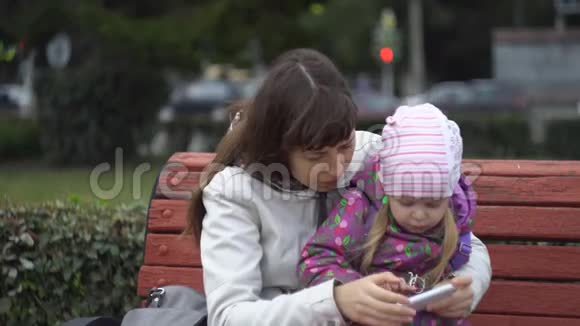 母女俩坐在户外的长凳上玩智能手机视频的预览图