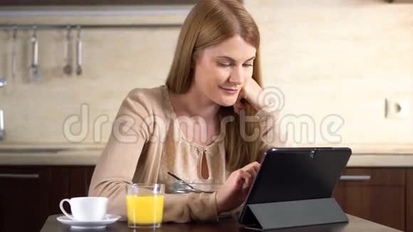 美丽的年轻迷人的女人早餐吃玉米片和使用她的平板电脑咖啡杯视频的预览图