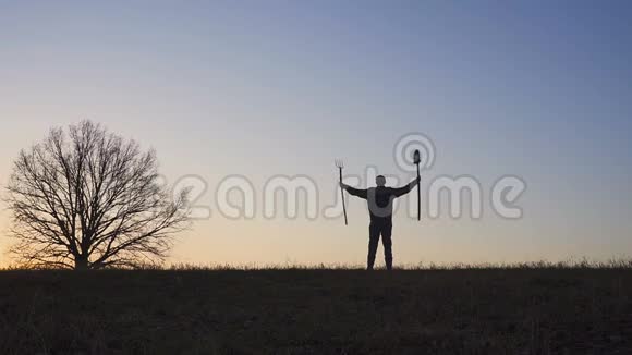 一个心满意足的农民用铲子和草叉挥动双手田野里日落或日出的剪影视频的预览图