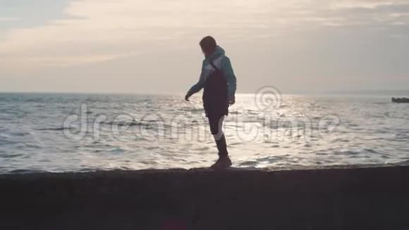 迷人的年轻女子穿着温暖的夹克在夕阳下的石滩上散步慢动作视频的预览图