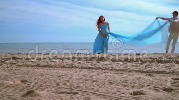 海滩上穿蓝色裙子的浪漫女人孕妇抱着肚子视频的预览图