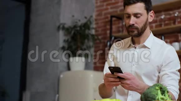 男人在家厨房用手机浏览英俊的年轻人在智能手机上浏览微笑着快乐一个男人吃视频的预览图