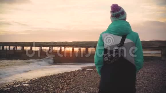 迷人的年轻女子穿着温暖的夹克在夕阳下的石滩上散步慢动作视频的预览图
