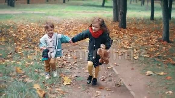 可爱的哥哥姐姐小男孩小女孩牵着手跑着拿着玩具穿过公园里的秋巷视频的预览图
