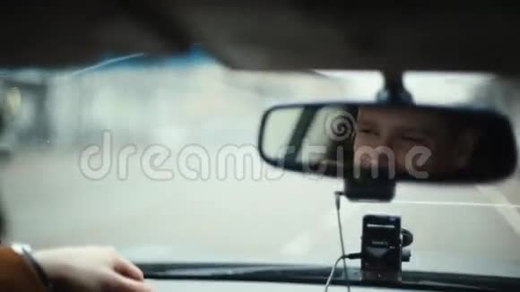 在城市里开车的时候对着镜子微笑的人英俊的男人的脸前窗上的雨滴开车旅行视频的预览图