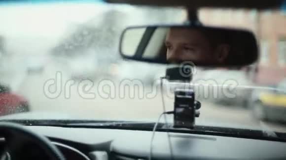 在城市里开车的时候对着镜子微笑的人英俊的男人的脸前窗上的雨滴开车旅行视频的预览图