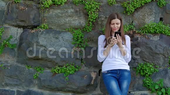 穿着白色衬衫的漂亮女孩用智能手机背景中野生石头的复古墙视频的预览图