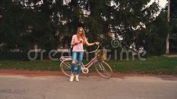 姜红头女孩坐在自行车上在夏季公园的智能手机上发短信穿着粉色衬衫和牛仔裤视频的预览图