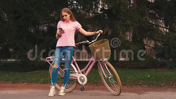 姜红头女孩坐在自行车上在夏季公园的智能手机上发短信穿着粉色衬衫和牛仔裤视频的预览图