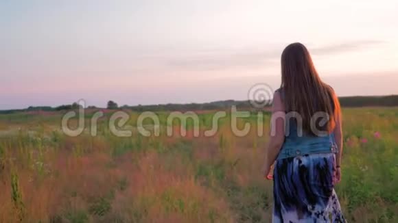 穿着裙子的年轻女子正沿着一片绿色的田野在野花丛中散步夕阳西下视频的预览图