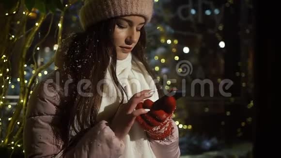美丽迷人的女孩站在一家装饰有花环的橱窗附近使用智能手机新年圣诞节视频的预览图