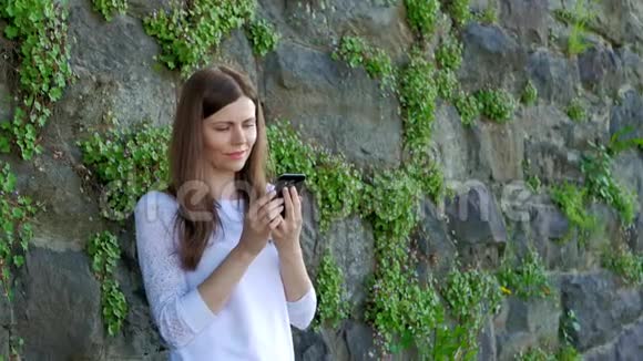 穿着白色衬衫的漂亮女孩用智能手机背景中野石的复古墙视频的预览图