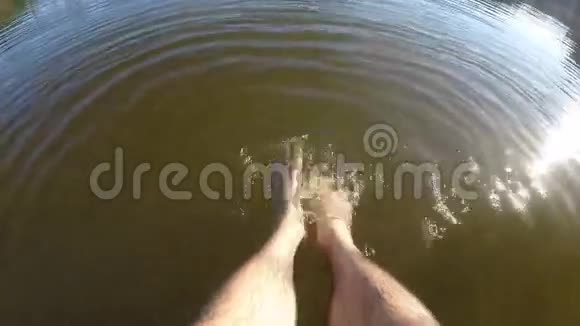 人在湖边或池塘边放松坐在木制码头的边缘在水中摆动一脚男腿视频的预览图