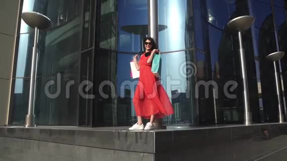 年轻漂亮的女孩在蓝色玻璃和倒影的建筑背景上穿着红色衣服拿着购物袋的布鲁内特视频的预览图